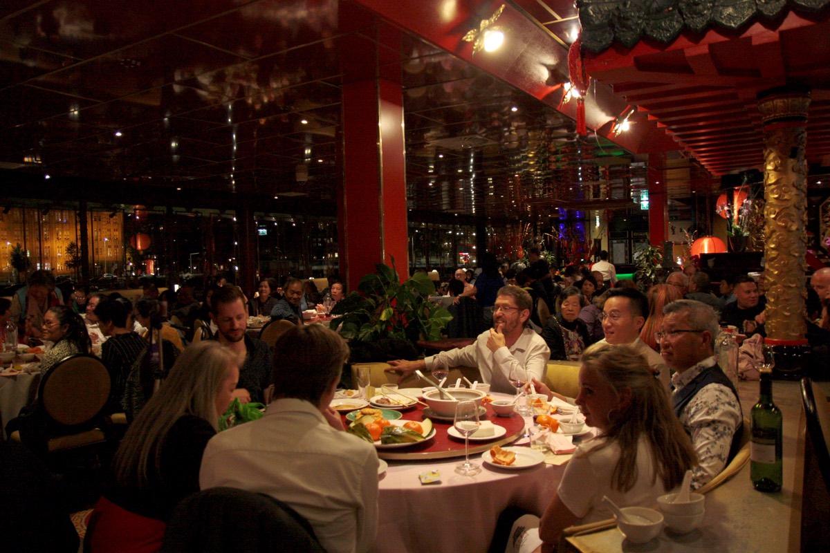 Dezelfde zaal later op de avond, aan tafel wordt gegeten en gepraat.