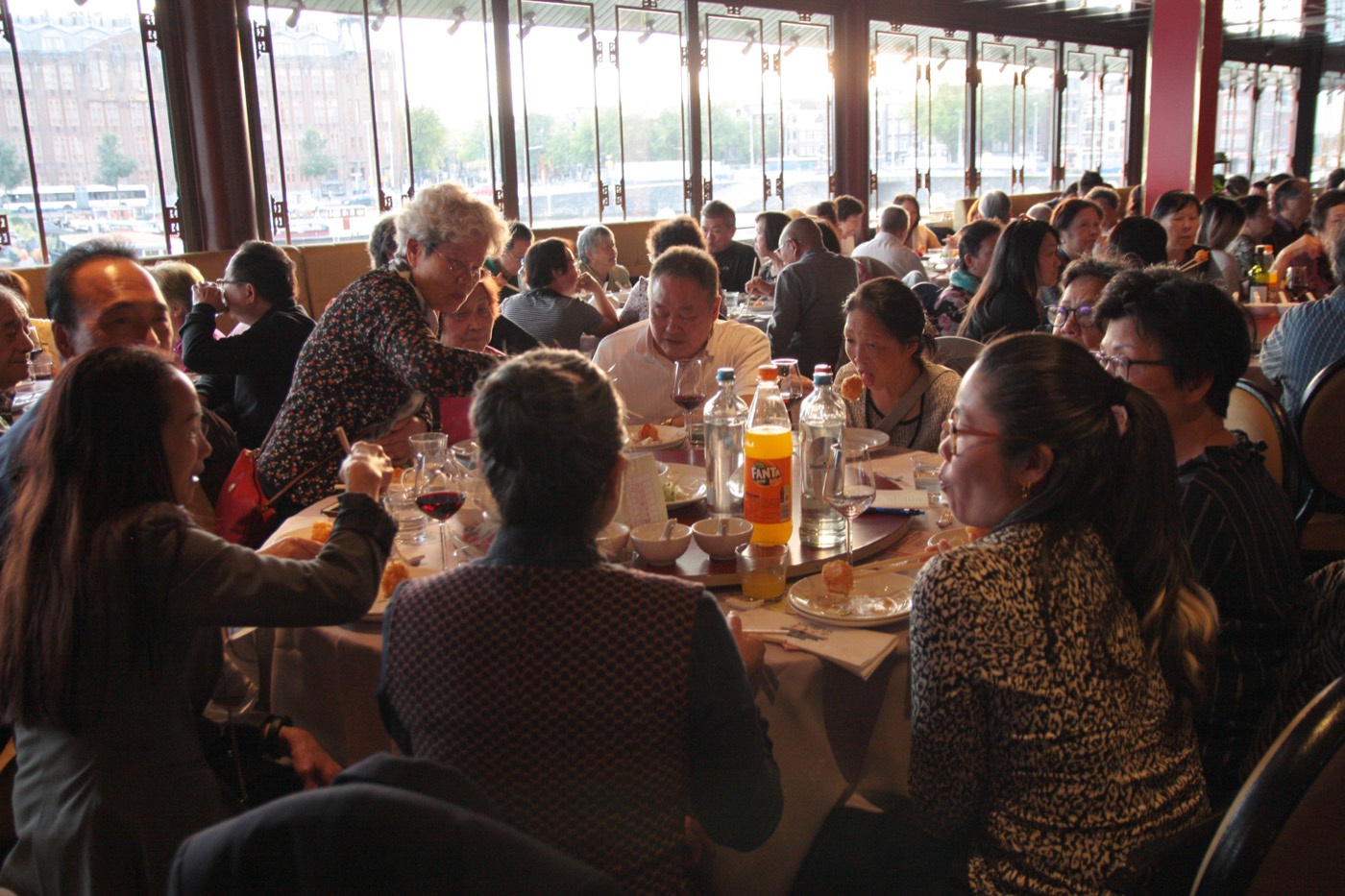 De volle zaal met ronde tafels, de ramen aan de kade erachter.